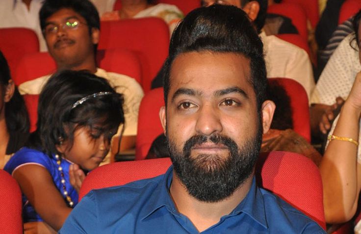 a man in blue shirt sitting in front of red chairs with other people behind him