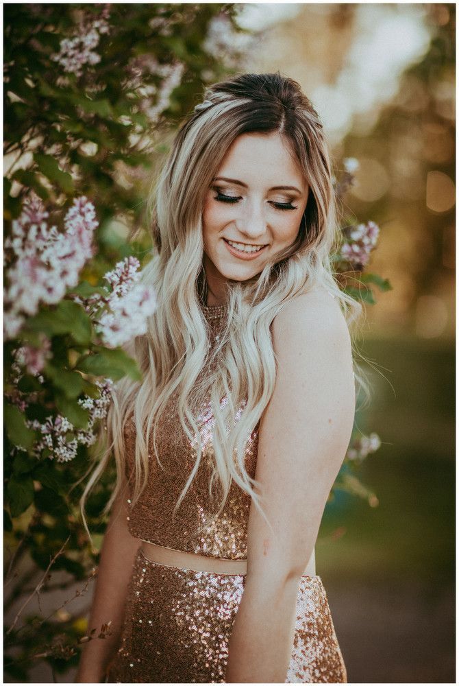 a woman with long blonde hair wearing a gold dress and standing in front of flowers