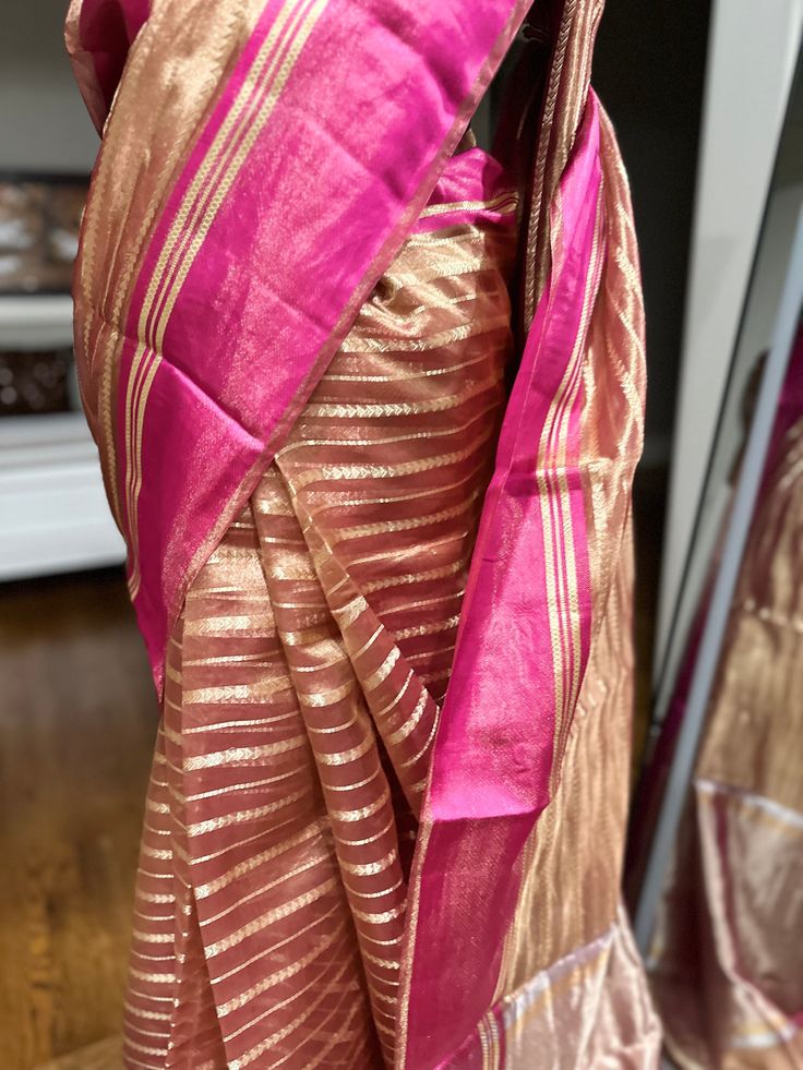 Indulge in ethereal elegance with our exquisite Kora Tissue Silk Saree, featuring a luxurious Katan Silk border adorned with shimmering gold zari. This masterpiece combines the delicate transparency of Kora tissue with the rich texture of Katan silk, creating a captivating contrast. The saree's ethereal beauty is further enhanced by the intricate gold zari work on the border, adding a touch of opulence. Perfect for weddings, cocktail parties, or any special occasion where you desire to make a lasting impression, this saree is a must-have for the discerning woman. Fabric: Kora by Tissue Silk Saree with Katan Silk Border Color: Rose Gold Embellishments: Gold Zari Occasion: Weddings, Parties, Festive Photos don't do justice .. this saree is just... glamorous Comes with fall/ Pico / tassels an Gold Cotton Silk Traditional Wear With Sheer Dupatta, Gold Traditional Wear With Sheer Dupatta In Cotton Silk, Gold Cotton Silk Saree For Party, Festive Gold Cotton Silk Traditional Wear, Party Gold Cotton Silk Saree, Gold Cotton Silk Dupatta For Party, Gold Cotton Silk Traditional Wear With Traditional Drape, Transitional Gold Chanderi Traditional Wear, Gold Chanderi Traditional Wear For Transitional Season