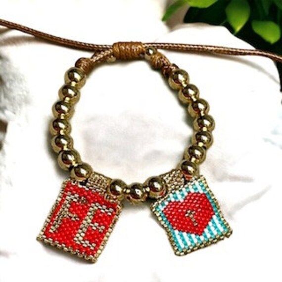 This beautiful and simple scapular bracelet is made of golden bronze tassels, leather and miyuki fabric with a double image.  *The first is the word "FE" (faith) and the second is the Cross of Christ inside a heart.  *This bracelet is adjustable so that you can wear it alone or in combination with other bracelets. *This is the perfect scapular bracelet to wear every day. It is a beautiful, traditional and unique religious jewelry that shows the signs of your faith. *The bracelet is made of leather and is adjustable to your wrist. PACKAGING: The jewelry is carefully packed in a soft peach skin bag. The peach skin bag is elegant and non-abrasive, which protects jewelry from scratches and damage during transport. This careful packaging helps keep jewelry in good condition and protected while Catholic Jewelry Scapular Leather Criss, Handmade Charm Bracelets Mother Mary, Brown Scapular, Brown Scapular Catholic, Multicolor Hand-strung Rosary Bracelet Gift, Catholic Bracelet, The Cross Of Christ, Catholic Jewelry, Protection Bracelet