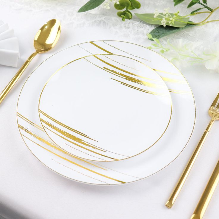 a white plate with gold lines on it next to two golden forks and spoons