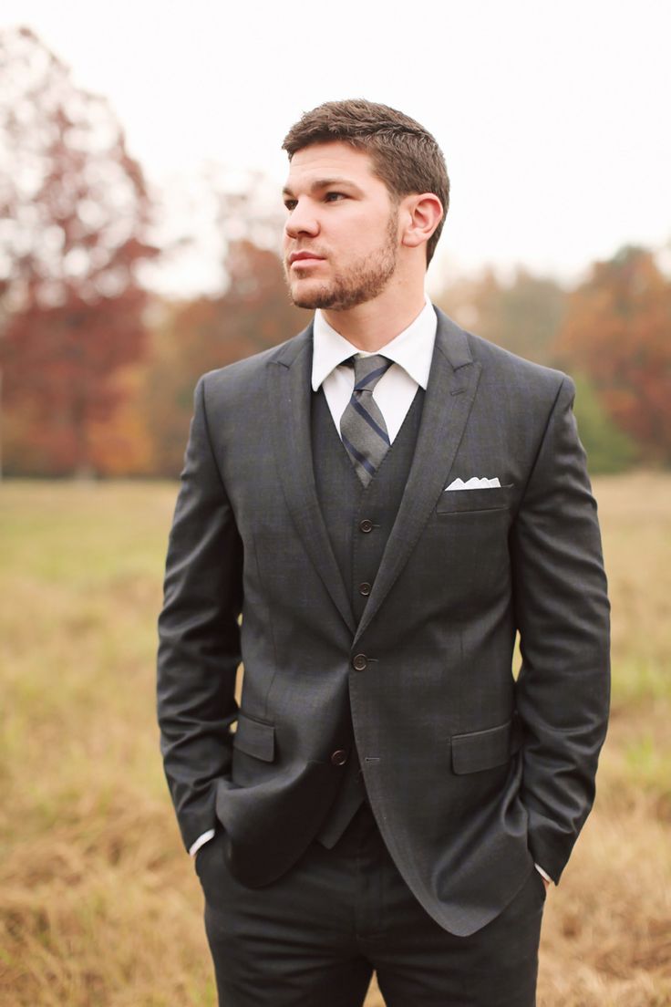 a man in a suit standing in a field with his hands in his pockets and looking off to the side