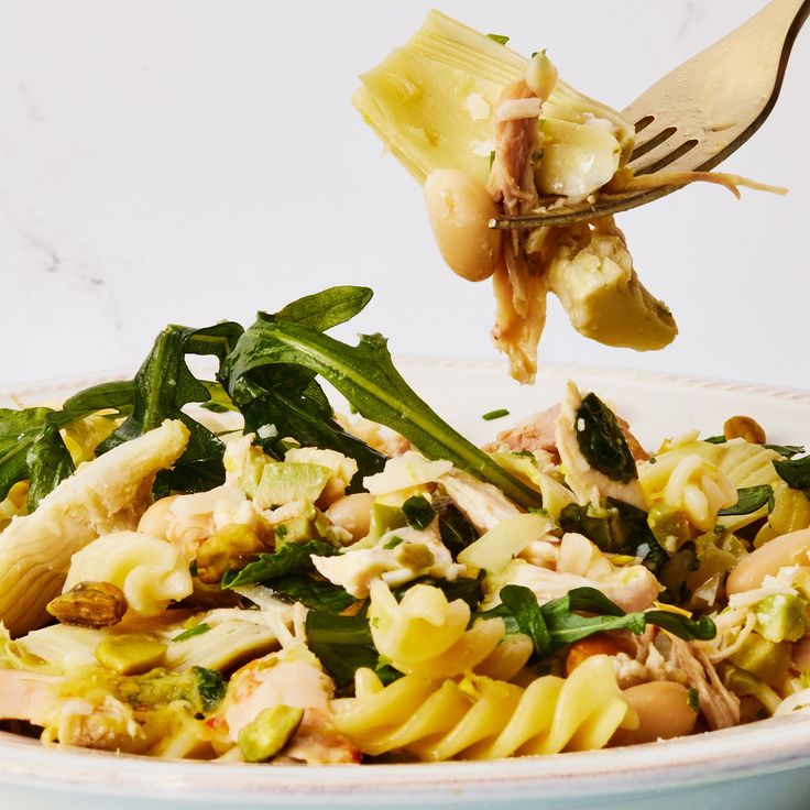 a fork is picking up some pasta from a white bowl filled with noodles and vegetables