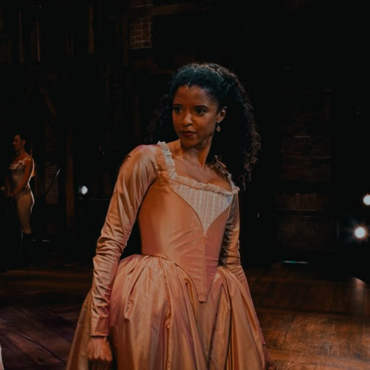 a woman in a pink dress standing on a wooden floor with other people behind her