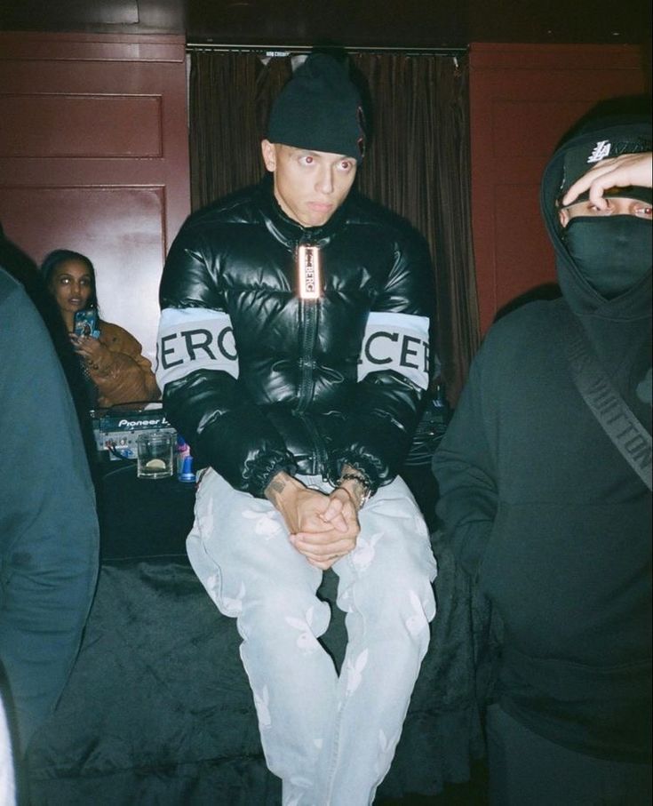 a man in a black jacket and hoodie sitting on a bed with other people behind him