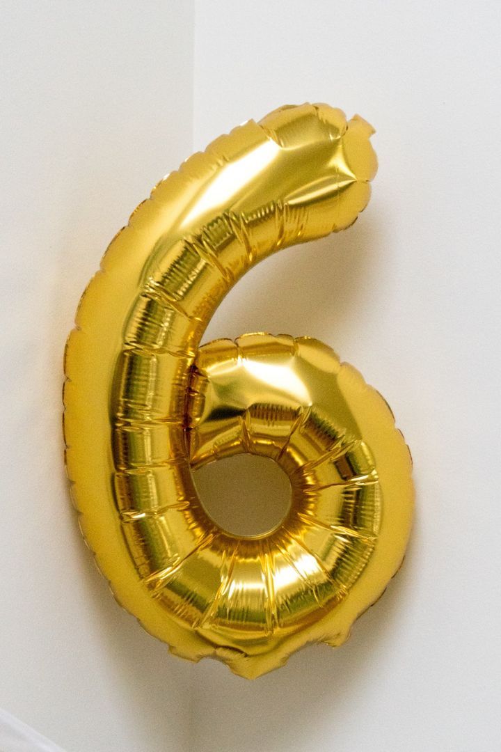 the number six is made out of gold foil balloons and sits on a white surface