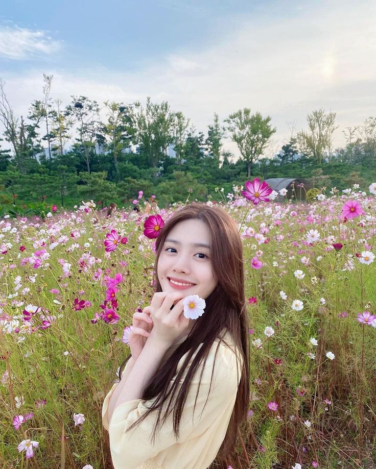 a young woman sitting in a field of flowers holding a flower up to her mouth