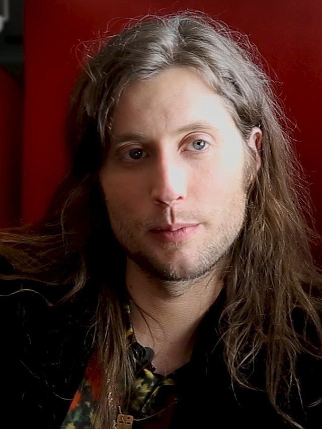 a man with long hair wearing a black jacket and looking at the camera while standing in front of a red wall