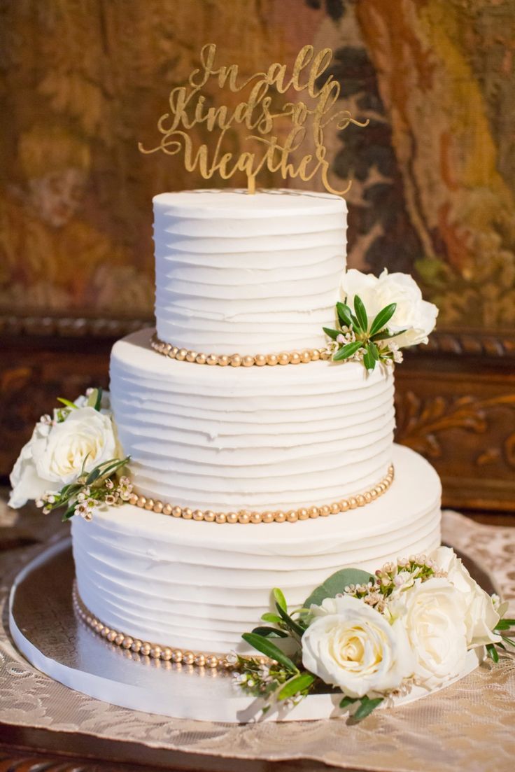 a white wedding cake with gold lettering on top and flowers in the bottom, sitting on a table