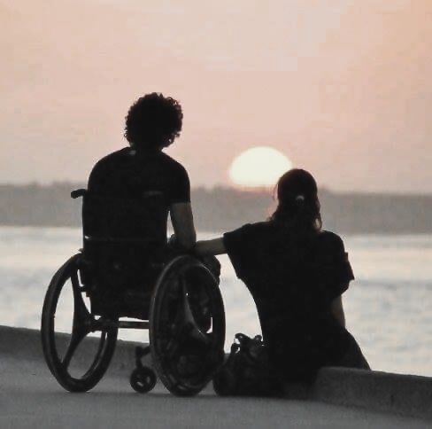 two people in wheelchairs sitting on the beach watching the sun go down over the water