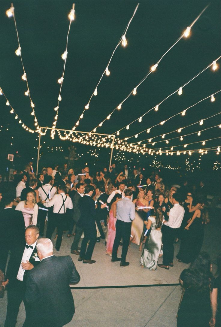 a group of people standing around each other on a dance floor with lights strung overhead