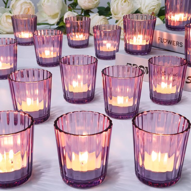 many purple glass cups with lit candles in them on a white table cloth next to flowers