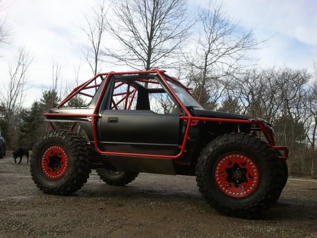 an off road vehicle with red rims parked in a parking lot next to trees