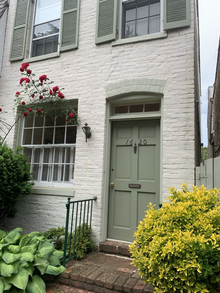 a white brick house with green shutters on the front and side doors, surrounded by flowers