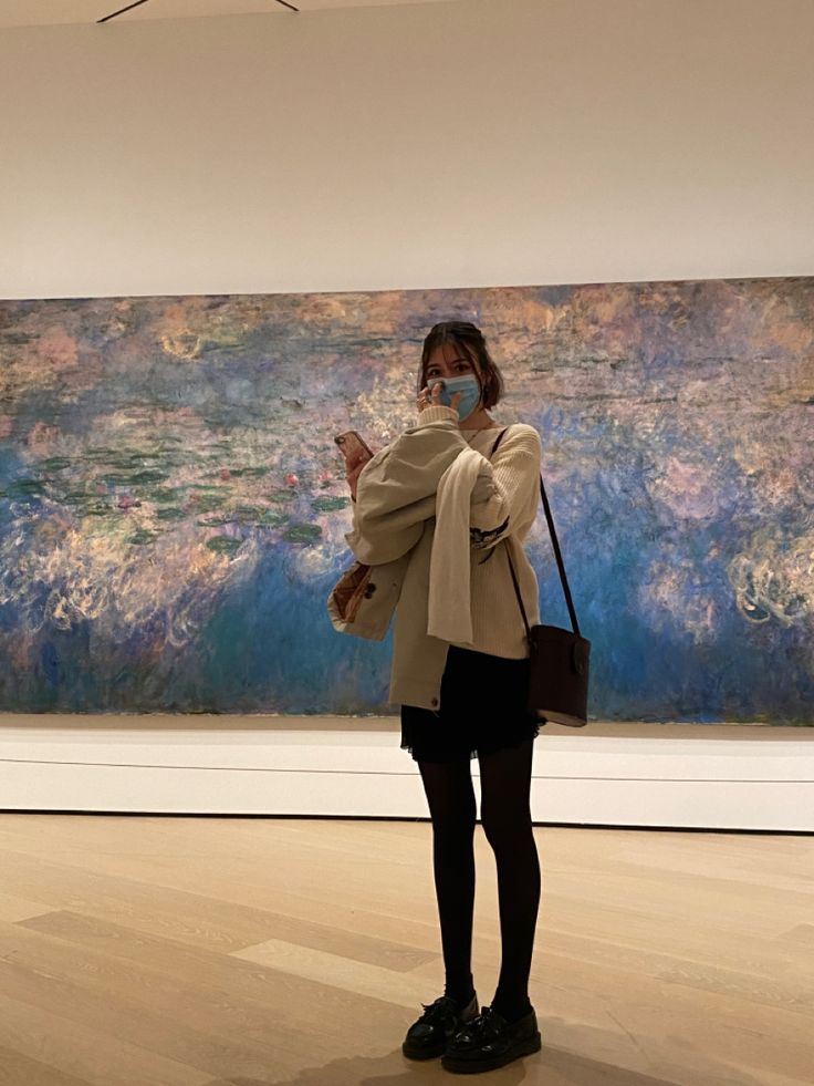 a woman standing in front of a painting wearing a face mask and looking at her cell phone