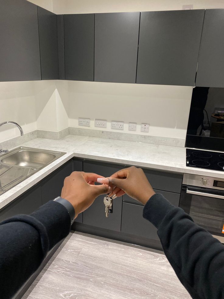 two people holding keys in a kitchen with grey cabinets and white counter tops, one person is reaching for the key