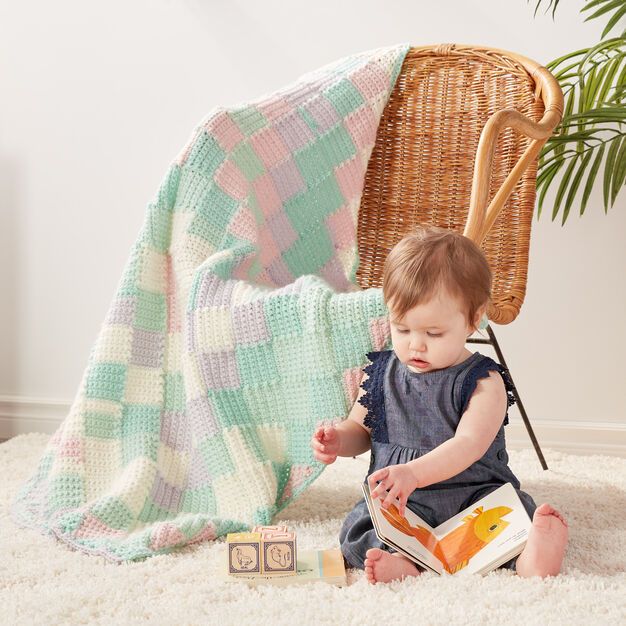 a baby sitting on the floor reading a book next to a blanket and chair with a potted plant