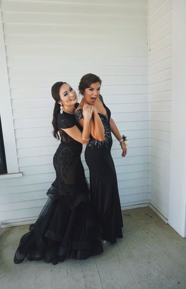 two women in black dresses standing next to each other on the porch with their arms around one another