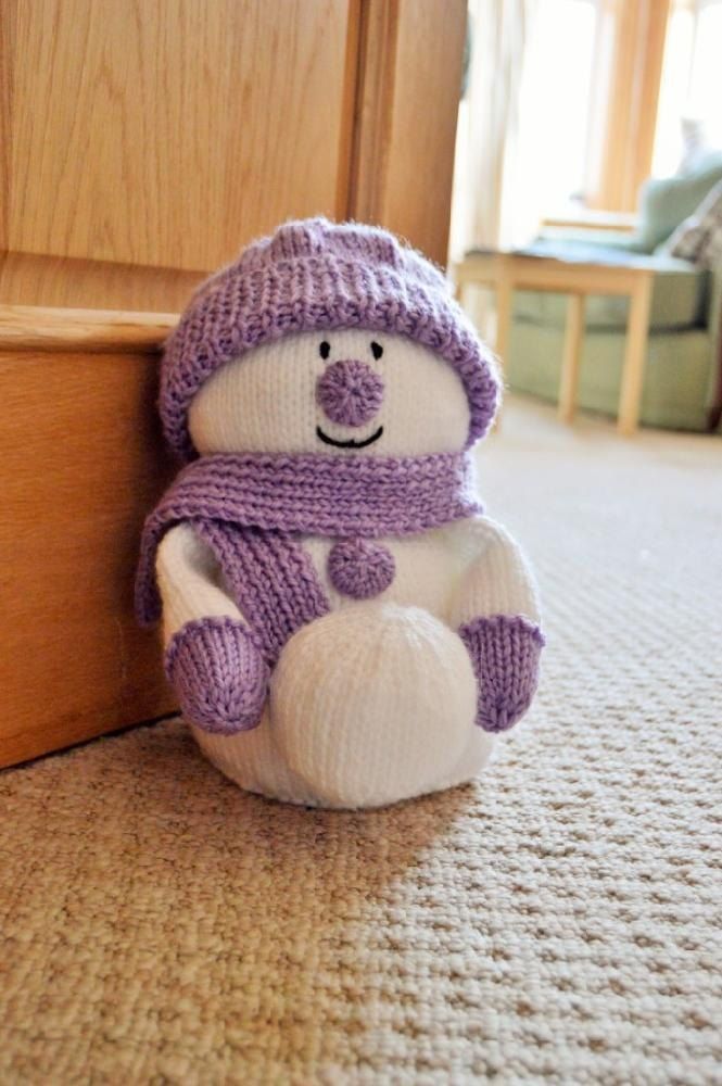 a stuffed snowman sitting on the floor next to a wooden door way and wearing a purple knitted hat