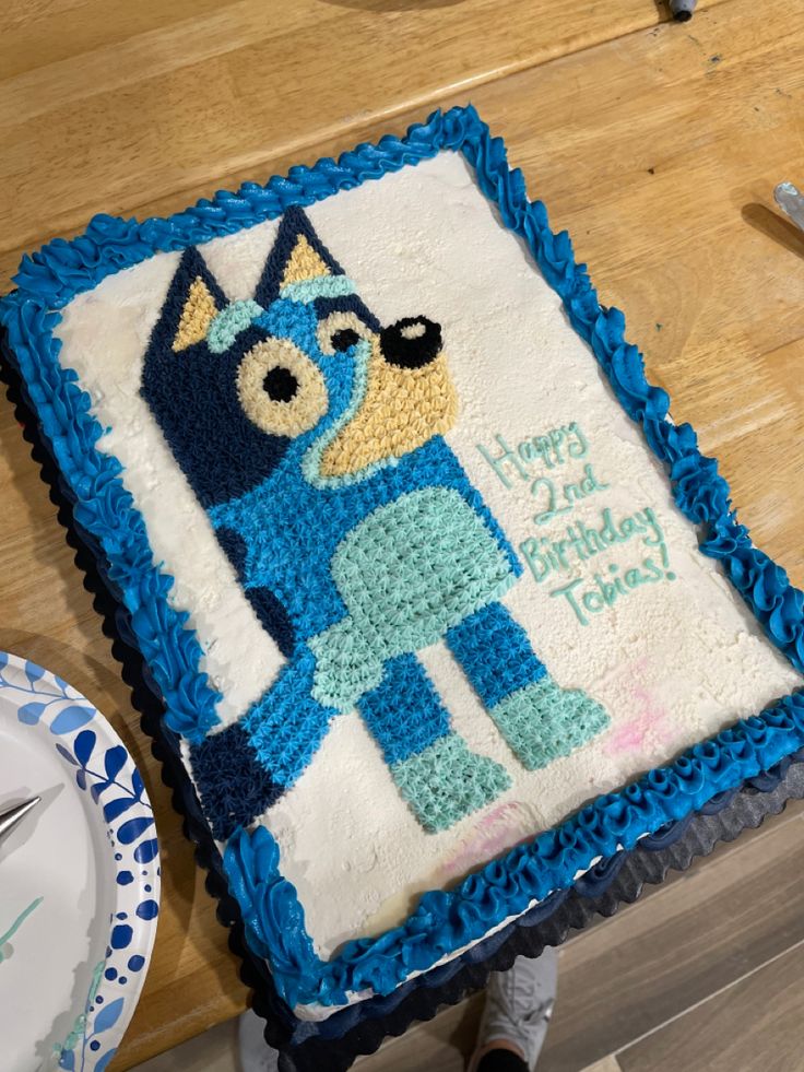 In this picture, we have Bluey the main character of the hit kids show ‘Bluey’ on a sheet cake. She is a blue heeler dog. Shes sky/baby blue in color with a tan snout, and navy spots. Theres a navy/ sky blue border around the cake along with a happy 2nd birthday text! Bluey Birthday Cake Rectangle, Bluey Cake Stencil, Bluey Cake Ideas Sheet Cake, Bluey Birthday Sheet Cake Ideas, Bluey Cookie Cake Ideas, Homemade Bluey Cake, Bluey 2nd Birthday Cake, Bluey Cake Ideas Easy, Bluey Cookie Cake