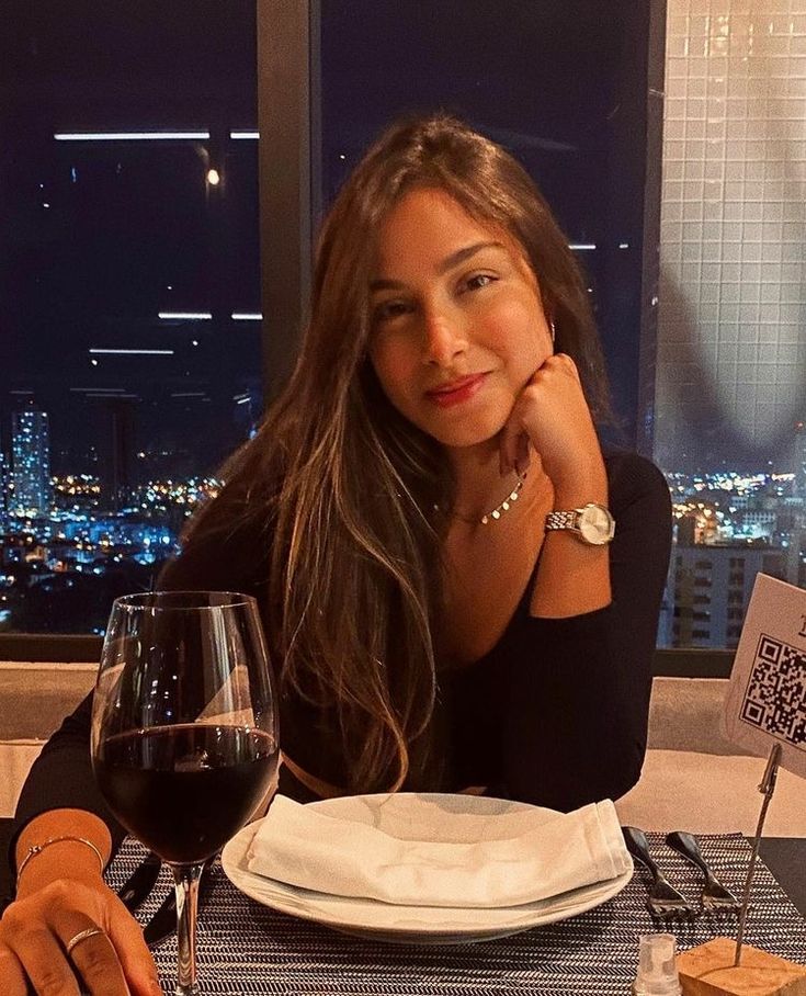 a woman sitting at a table with a glass of wine in front of her and the city lights behind her