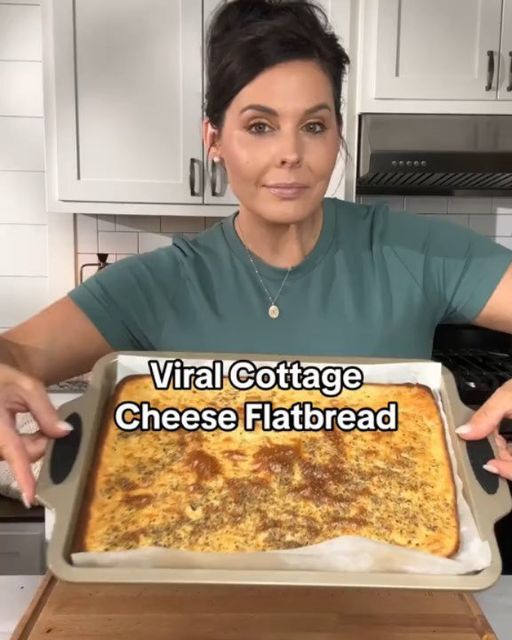 a woman holding up a large tray of cheese flatbread in her kitchen with the words virtual cottage cheese flatbread on it