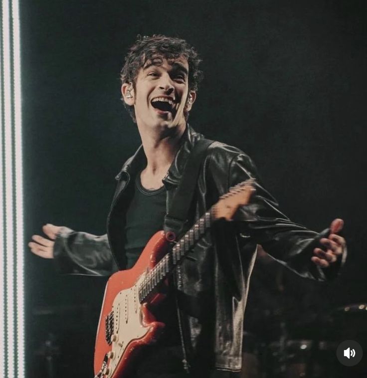 a man holding a red guitar in his right hand and singing into the microphone on stage