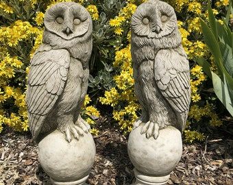 two statues of owls sitting on top of each other in front of flowers and bushes