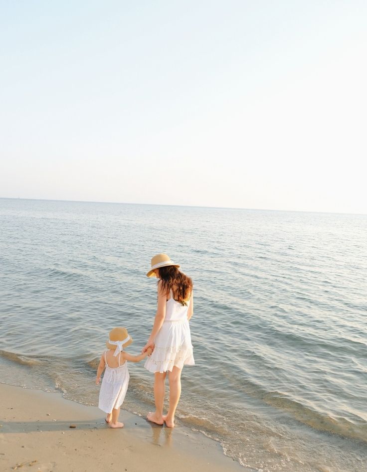 a mother and daughter on the beach holding hands while standing in front of the water