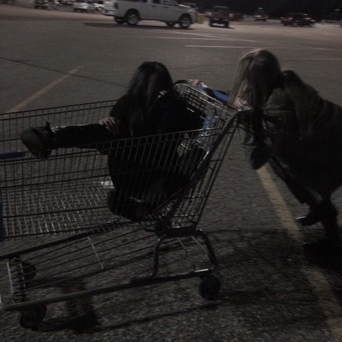 two people pushing a shopping cart in an empty parking lot at night, with the lights on