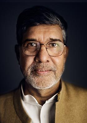 a man with glasses is posing for a photo in front of a dark background, wearing a brown jacket and white shirt