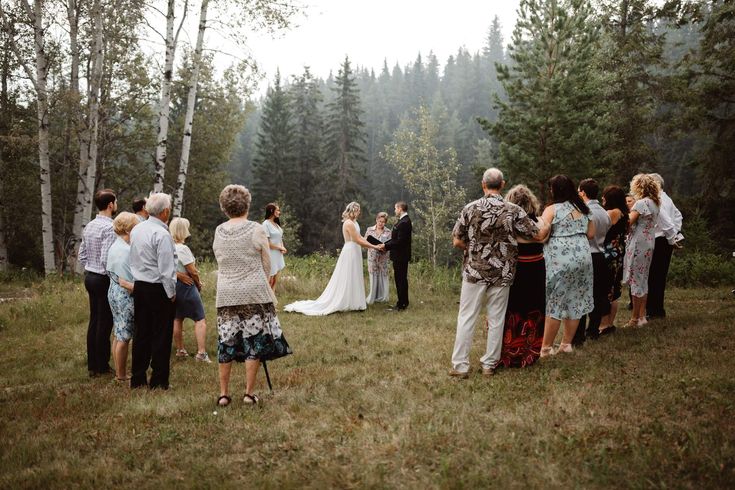 a group of people standing around each other in the grass with trees and bushes behind them