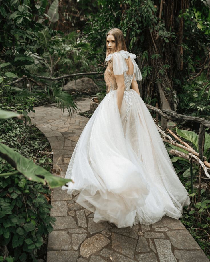 a woman in a white wedding dress walking through the woods on a stone path with greenery behind her