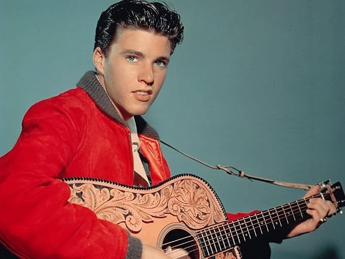 elvis presley holding an acoustic guitar in his right hand and wearing a red jacket with paisley designs on it