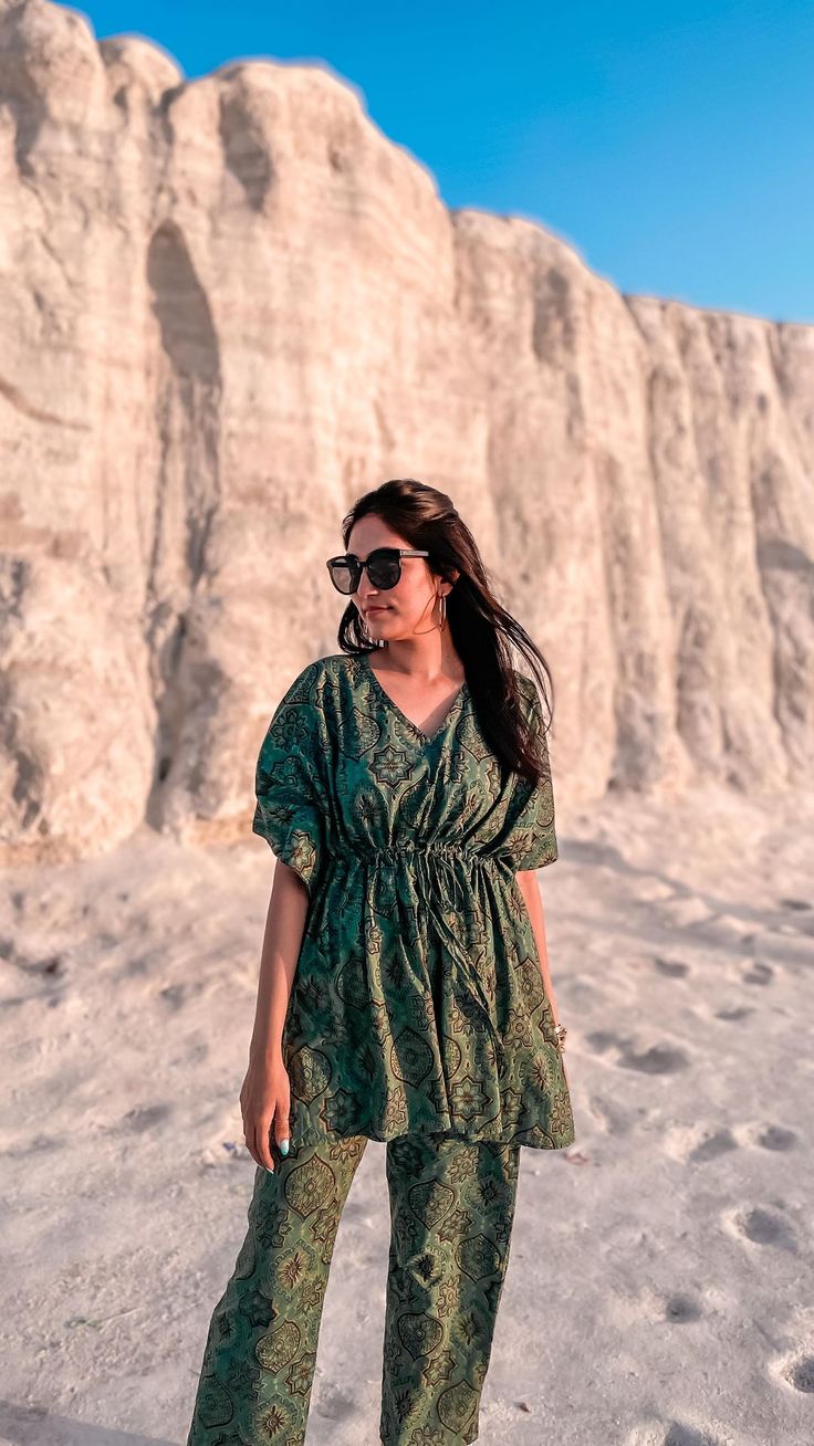 a woman standing in the sand wearing sunglasses and a green top with an animal print on it