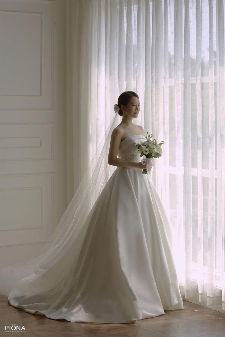 a woman standing in front of a window wearing a wedding dress and holding a bouquet