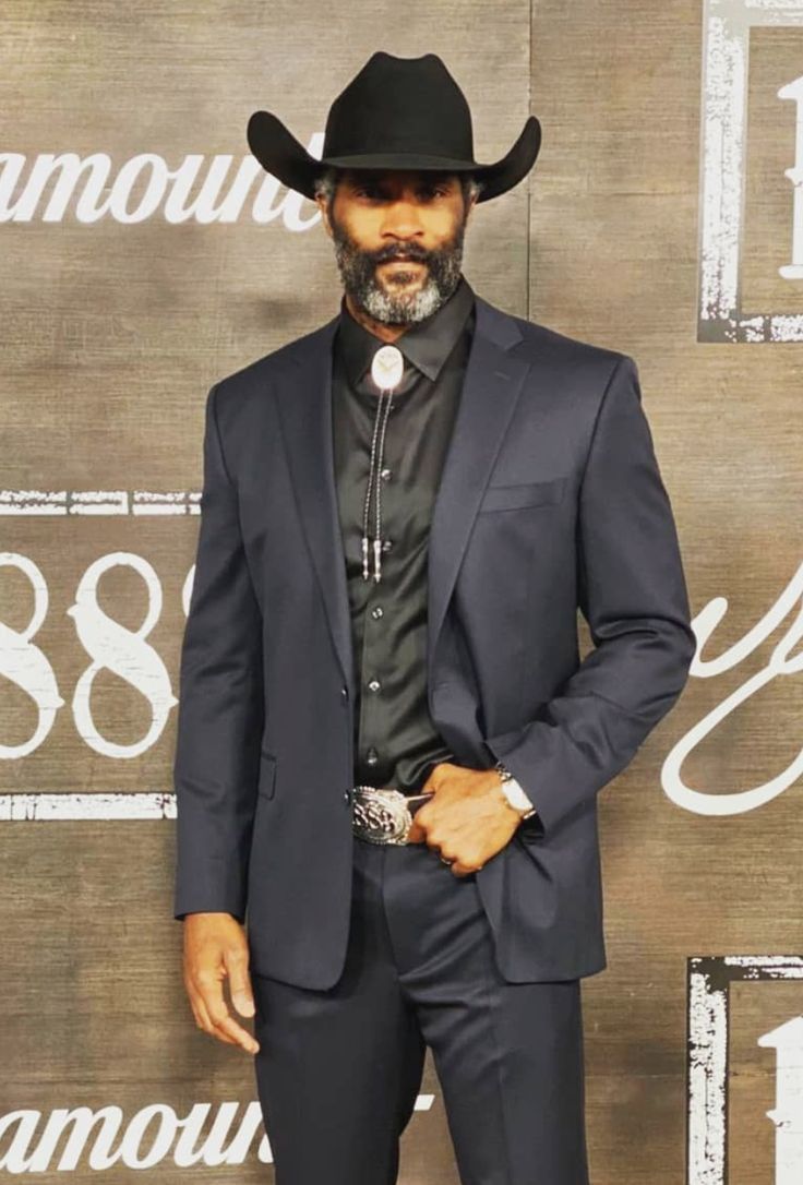 a man in a black suit and cowboy hat poses for a photo on the red carpet