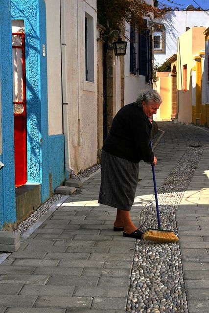 an old woman sweeping up the sidewalk with a broom in front of colorful buildings and cobblestone streets