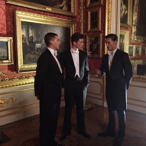 three men in suits standing next to each other looking at paintings on the wall behind them