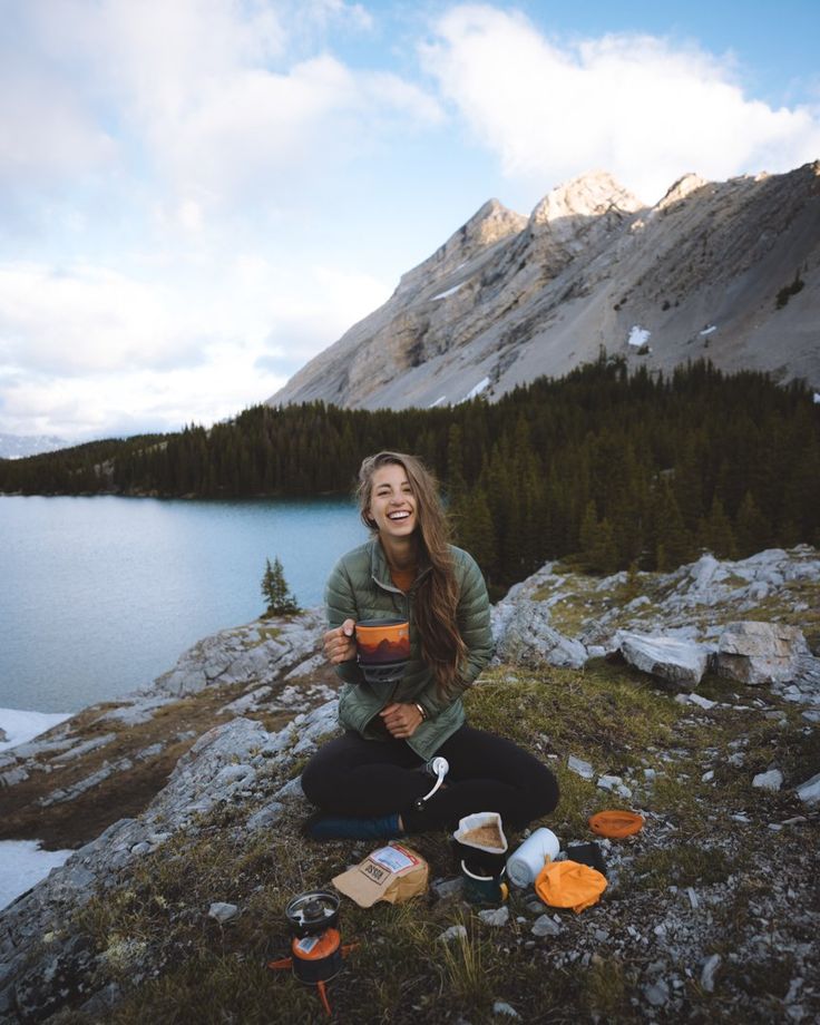 a woman sitting on top of a mountain next to a lake holding an orange cup