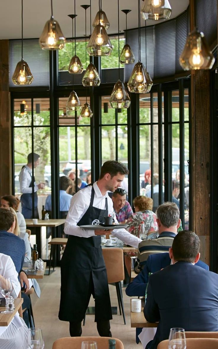 a waiter is standing in front of a group of diners at a restaurant with lights hanging from the ceiling