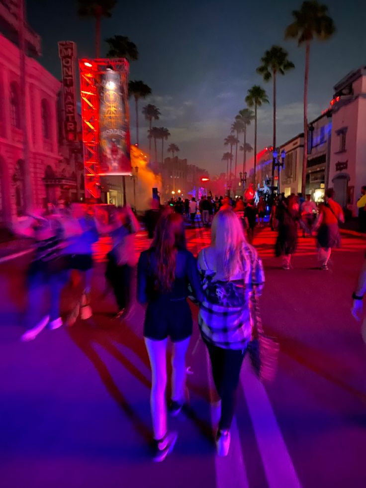 people walking down the street at night with neon lights and palm trees in the background