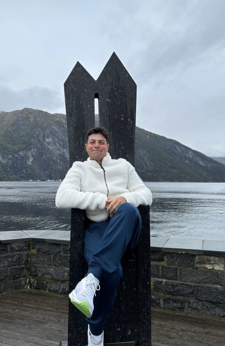 a man sitting on top of a wooden chair next to the ocean with mountains in the background