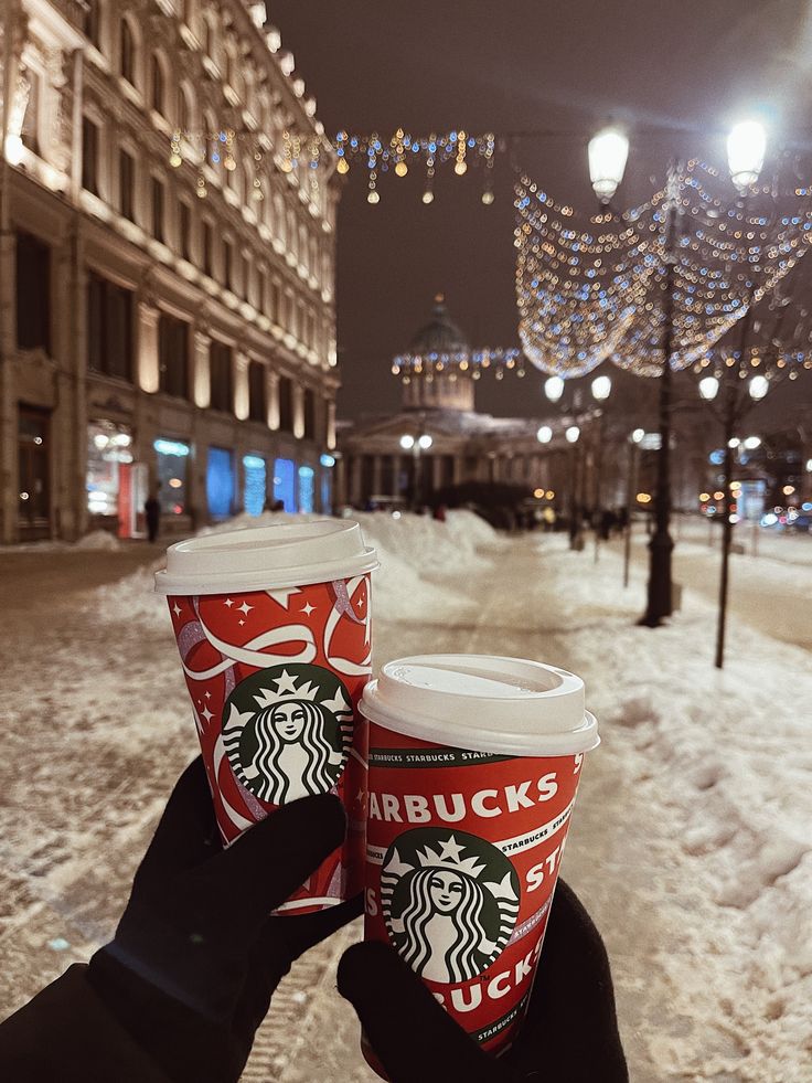 two cups of starbucks coffee are held up in front of a snowy street at night