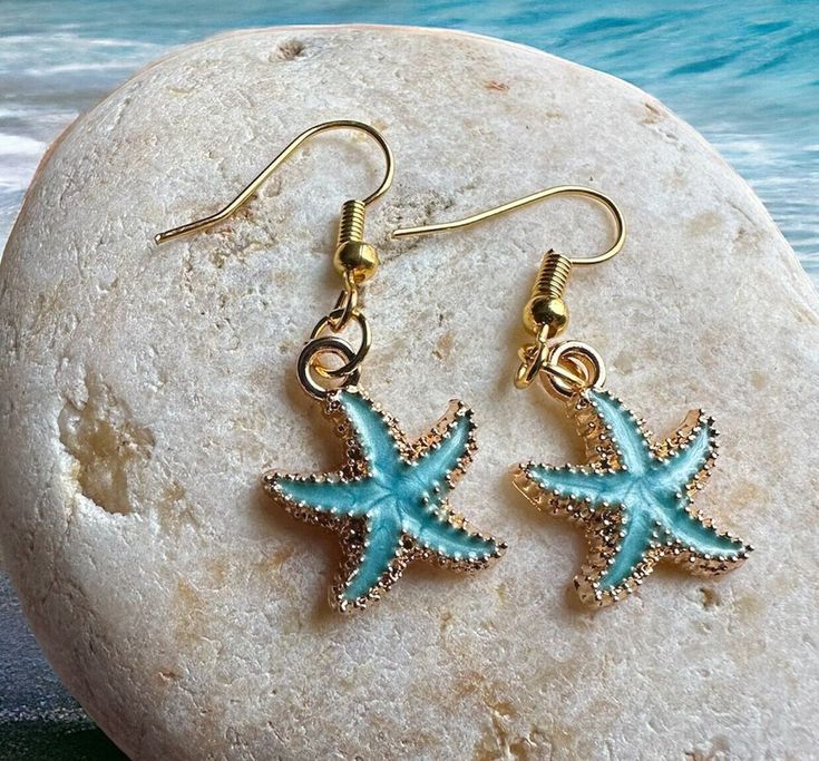 "These stylish starfish earrings will transport you right to the beach.  With dainty gold accents and gorgeous Caribbean blue colors, these beachy earrings perfectly capture the unparalleled beauty of the ocean. Our dainty sea star earrings measure approximately 1 1/4\" in length - lightweight and comfortable for everyday wear.   All of our ocean-inspired jewelry radiates the warmth and shimmer of the sea and these beautiful blue starfish earring are no exception. Ideal for beach lovers or marin Blue Star Jewelry For Vacation, Blue Star Shaped Jewelry For Vacation, Blue Starfish Charm Jewelry For Beach Season, Blue Jewelry With Starfish Charm For Beach Season, Ocean-inspired Starfish Charm Earrings, Nickel-free Starfish Earrings Ocean-inspired, Blue Starfish Charm Earrings For Gift, Starfish Charm Earrings Gift, Starfish Charm Earrings For Gift