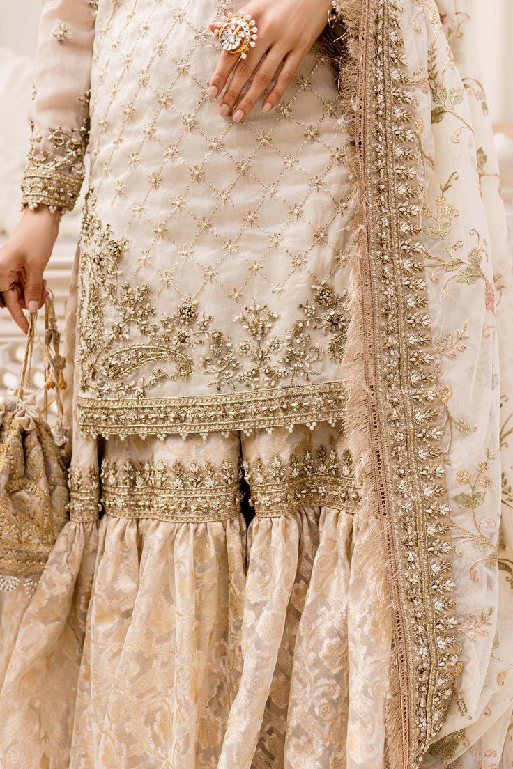 Pakistani Bridal Dress in Kameez and Gharara Style is a breathtaking masterpiece that will give you a traditional look on the big day. Lavish details of Kundan, zari, gotta, and Kiran work makes this Gharara Dress an epitome of beauty and your foremost priority. Bridal Kameez: The kameez has an alluring ivory gold shade and it is embellished with hand-crafted details of gota and zari work. Kundan, pearls, sequins, and dabka give a traditional touch to this kameez, making it a perfect choice to p Bridal Gharara, Gharara Designs, Pakistani Bridal Dress, Desi Wedding Dresses, Latest Bridal Dresses, Bridal Dresses Pakistan, Pakistani Fancy Dresses, Bridal Dress Fashion, Maria B