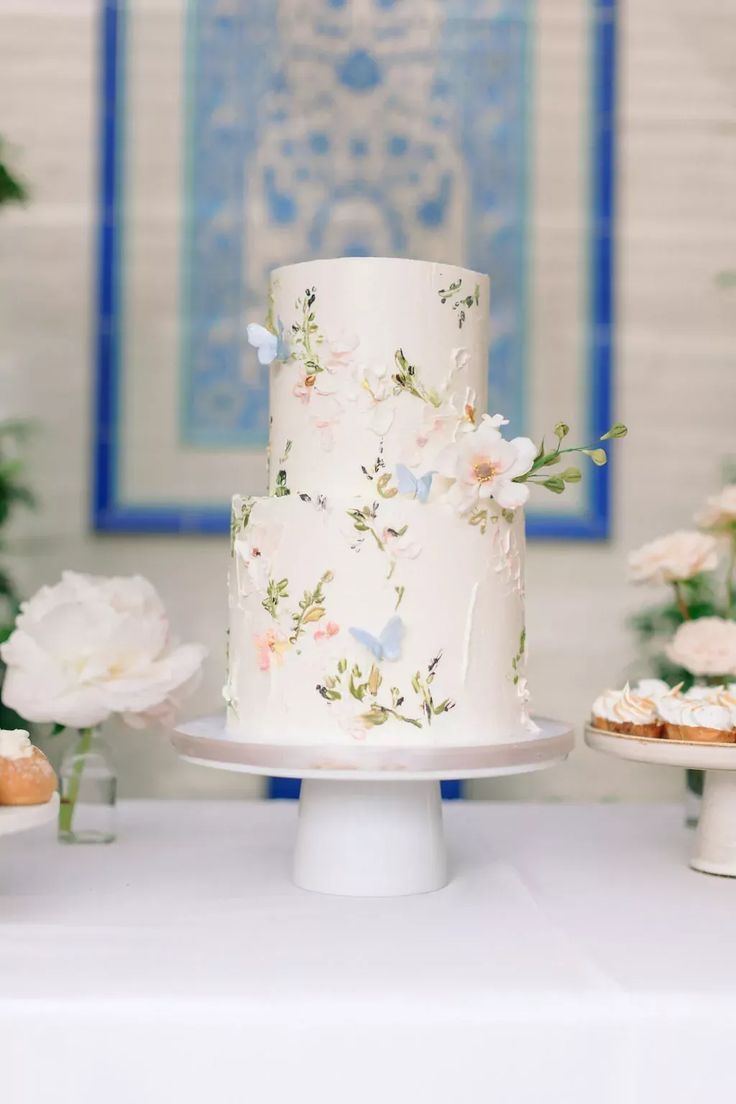 a white wedding cake with flowers on the top is sitting on a table next to cupcakes