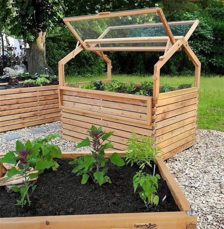 a wooden garden box with plants growing in it on top of graveled ground next to grass and trees