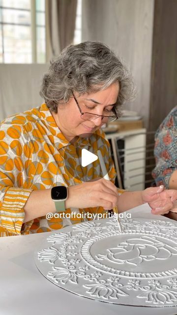 an older woman sitting at a table working on a piece of art with another woman in the background