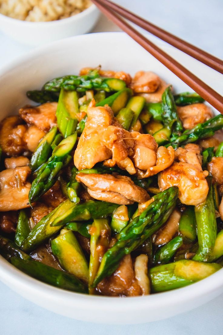 a bowl filled with chicken and asparagus next to chopsticks on a table